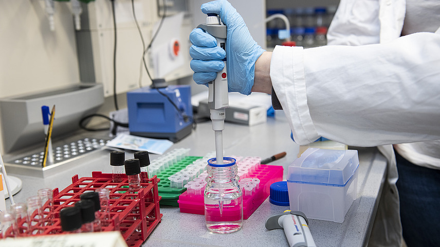 Two people in white coats are standing in a laboratory with only their hands visible. One of them is pointing an electric pipette into a glass container.