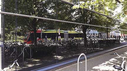 A reflective glass façade shows a street on which a colorful bus and a row of bicycles are parked. Trees provide shade and are visible in the reflection of the glass. Passers-by and other buildings can be seen in the background