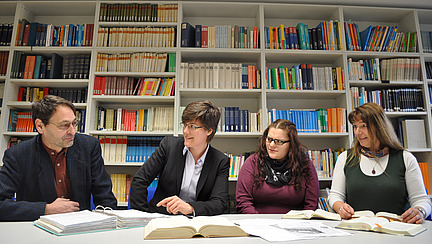 Four people sit at a table in front of a wall of books with books and notes in front of them