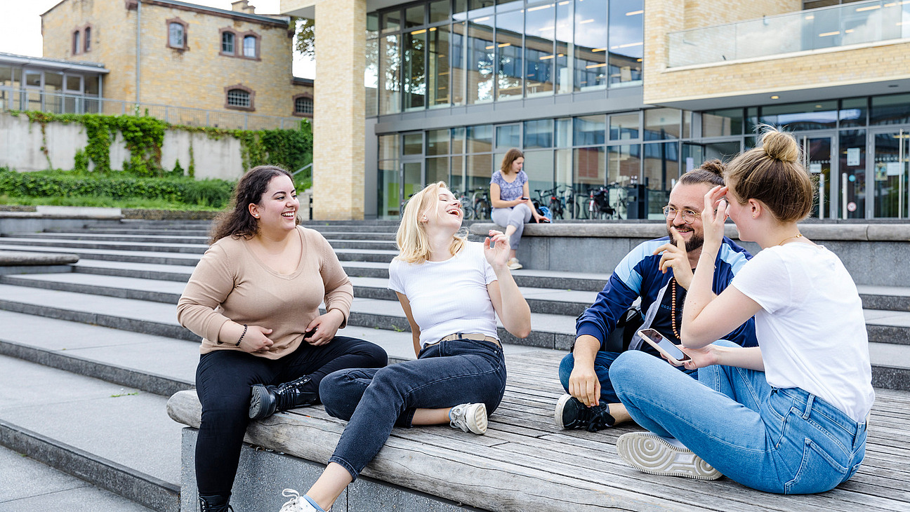 The picture shows a group of five people sitting together on a wooden bench and communicating with each other. They are laughing and seem to be having a good time. In the background are modern buildings and some people who are staying there. The scene conveys a relaxed and friendly atmosphere.