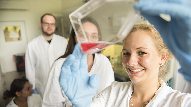 Three people in white coats. A smiling woman holds a plastic container with red liquid towards the viewer.