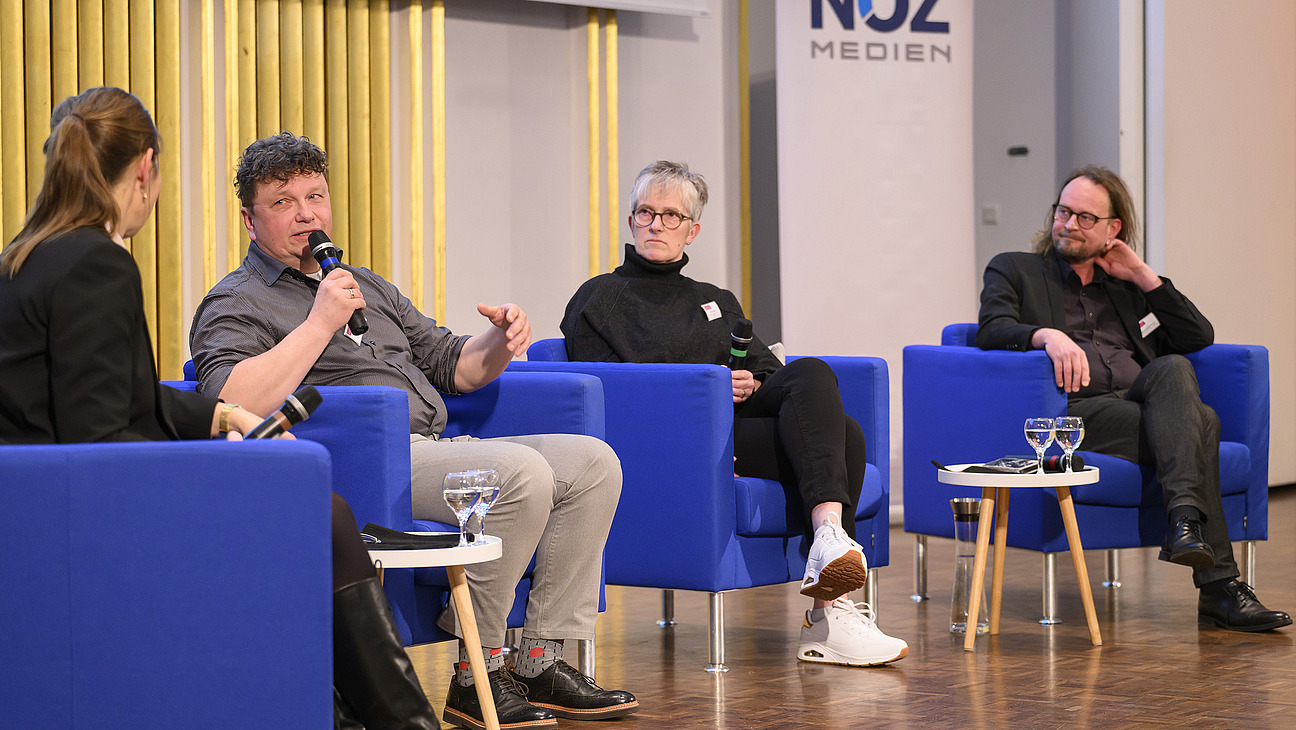 A man speaks on stage while three other people listen.