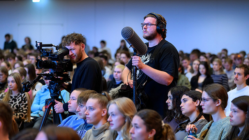 Ein Kameramann und ein Tonmann stehen in einem Saal zwischen Zuschauerreihen