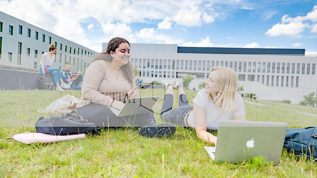 Zwei Studentinnen auf dem begrünten Platz neben der Bibliothek. Eine von ihnen sitzt, die andere liegt auf dem Rasen, beide haben Laptops vor sich