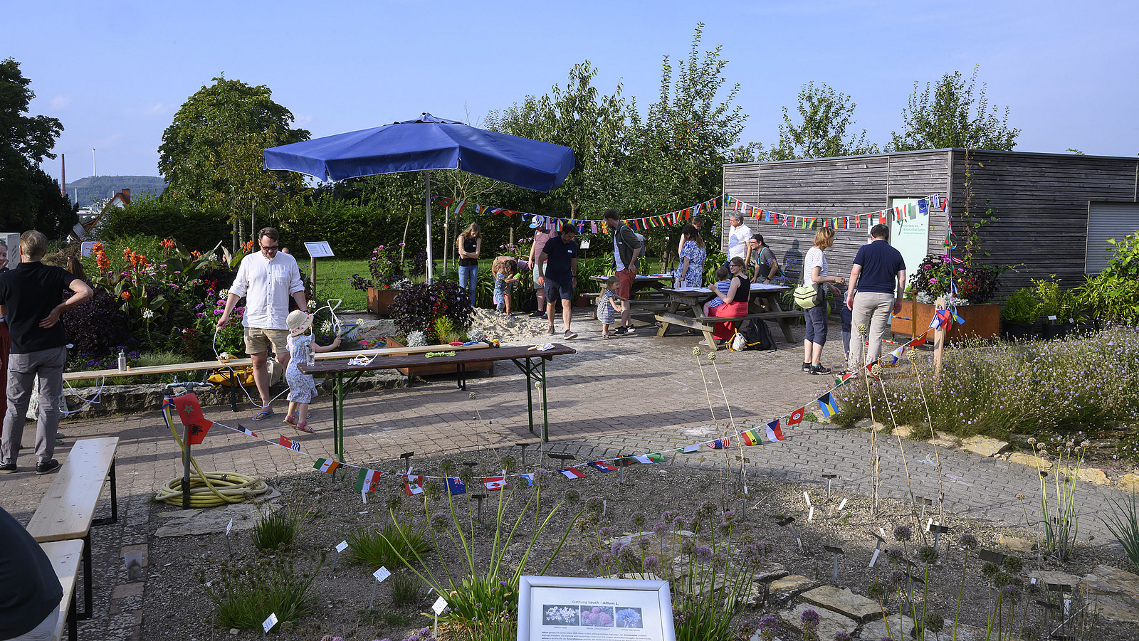 A garden with various surfaces, a parasol in the background, a wooden building, colorful flags and a few people.