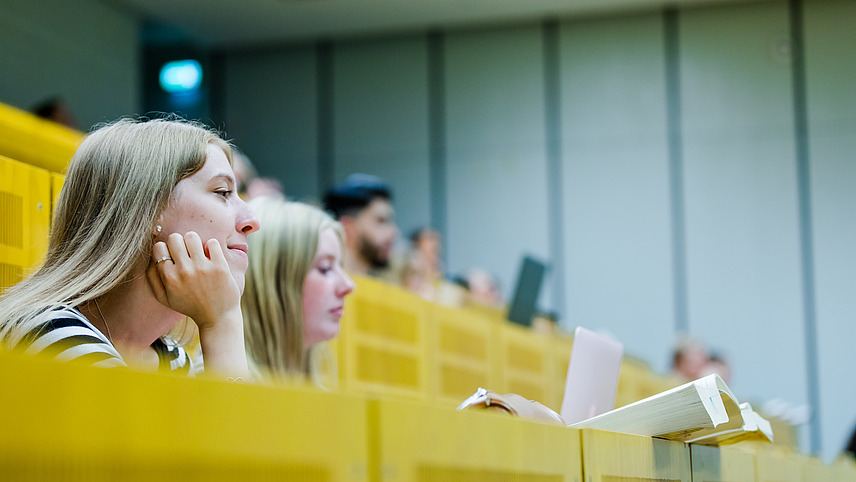 Zwei Studentinnen in einem Hörsaal. Ihre Bücher ragen über die Lehne der Sitzplätze vor ihnen.