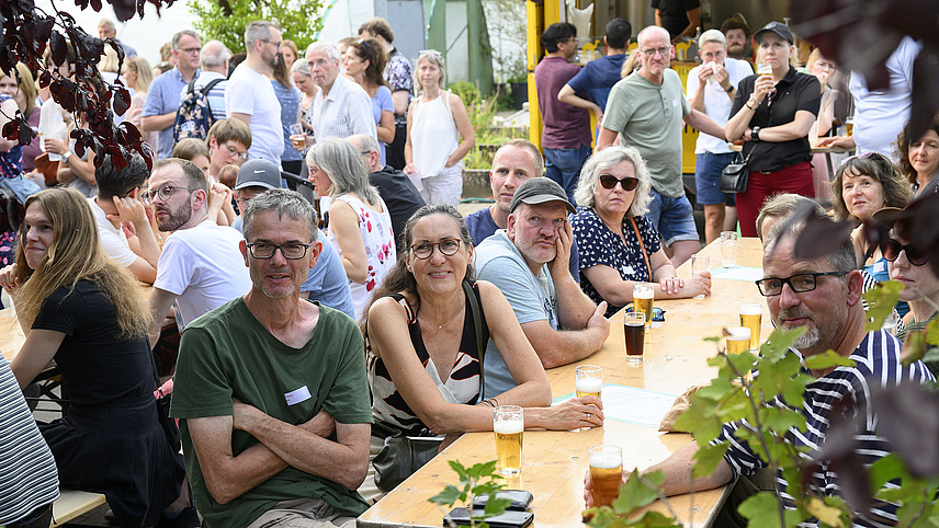 Mehrere fröhliche Personen sitzen mit Getränken bei einer Gartenparty.