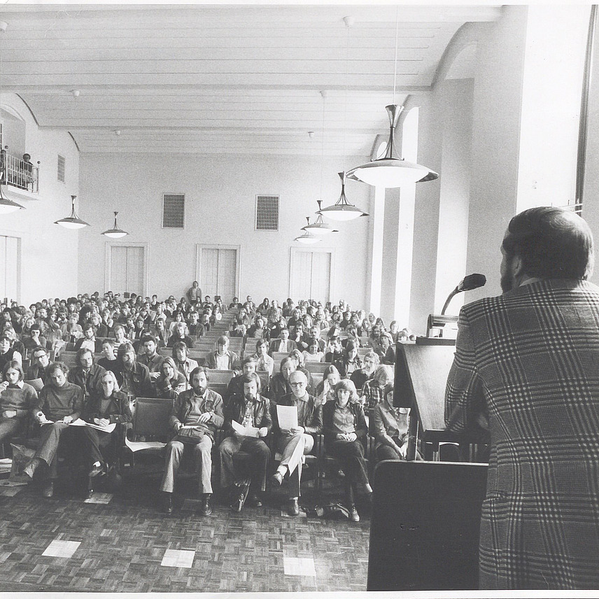 A black and white shot of a lecture in a large hall. In the foreground, a speaker stands at a lectern while many members of the audience sit in rows and look towards him. 