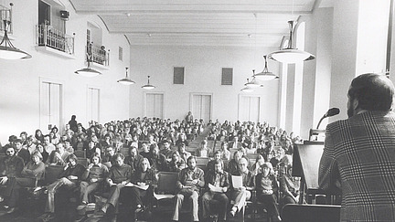 Eine Schwarz-Weiß-Aufnahme eines Vortrags in einem großen Saal. Im Vordergrund steht ein Redner an einem Pult, während viele Zuhörer in Reihen sitzen und auf ihn blicken.