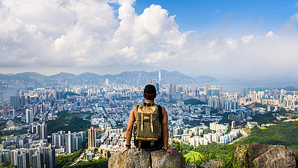 A young man stands on a mountain with a rucksack and looks at the town in the valley in front of him, right by the sea.