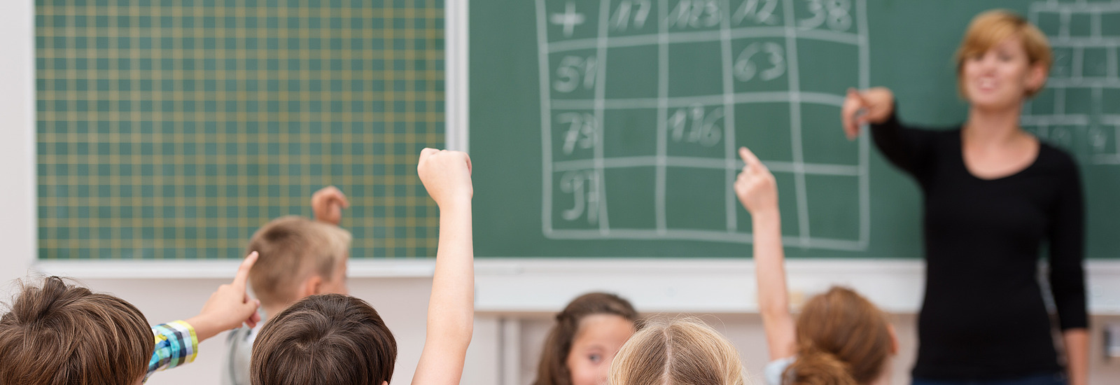 In einem Klassenzimmer sitzen mehrere Kinder an ihren Tischen, die Hände erhoben, um sich zu melden. Ein Junge in einem karierten Hemd und ein Mädchen mit einem rosa T-Shirt sind gut sichtbar. Im Vordergrund schaut die Lehrerin, die an einer Tafel steht und anspricht, in die Richtung der Schüler.