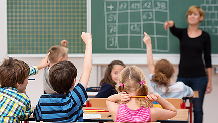 Several children are sitting at their desks in a classroom, hands raised to sign in. A boy in a checked shirt and a girl in a pink T-shirt are clearly visible. In the foreground, the teacher, who is standing at a blackboard and addressing the pupils, looks in their direction.