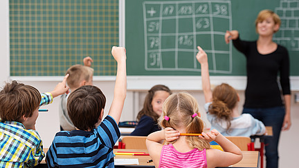 In einem Klassenzimmer sitzen mehrere Kinder an ihren Tischen, die Hände erhoben, um sich zu melden. Ein Junge in einem karierten Hemd und ein Mädchen mit einem rosa T-Shirt sind gut sichtbar. Im Vordergrund schaut die Lehrerin, die an einer Tafel steht und anspricht, in die Richtung der Schüler.