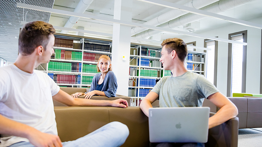 Three young people are sitting on a comfortable sofa in a modern room. Two of them are looking at each other and appear to be having a conversation, while one of them has a laptop on his knees. Colorful shelves with books or other materials can be seen in the background