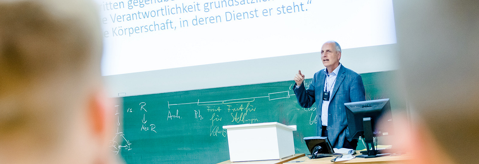 Looking past the heads of two students. A lecturer is giving a lecture. In the background is a fully written blackboard and a presentation is projected onto the wall above it.