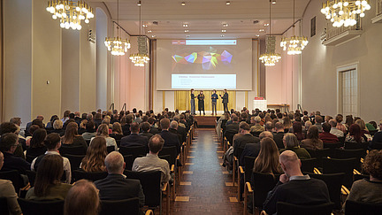 A choir is singing on stage in a large hall. Many people are sitting in the audience.