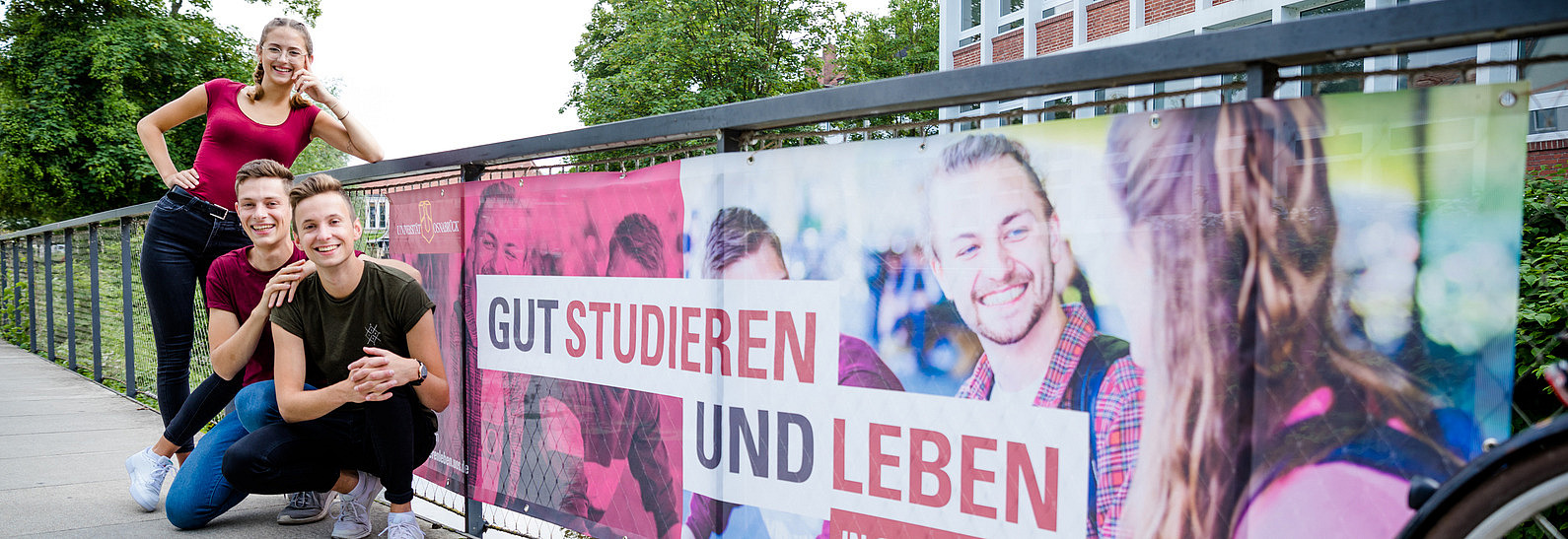 Drei Studierende posieren vor einem Geländer an dem ein Werbeschild mit der Aufschrift "Gut studieren und leben" hängt.