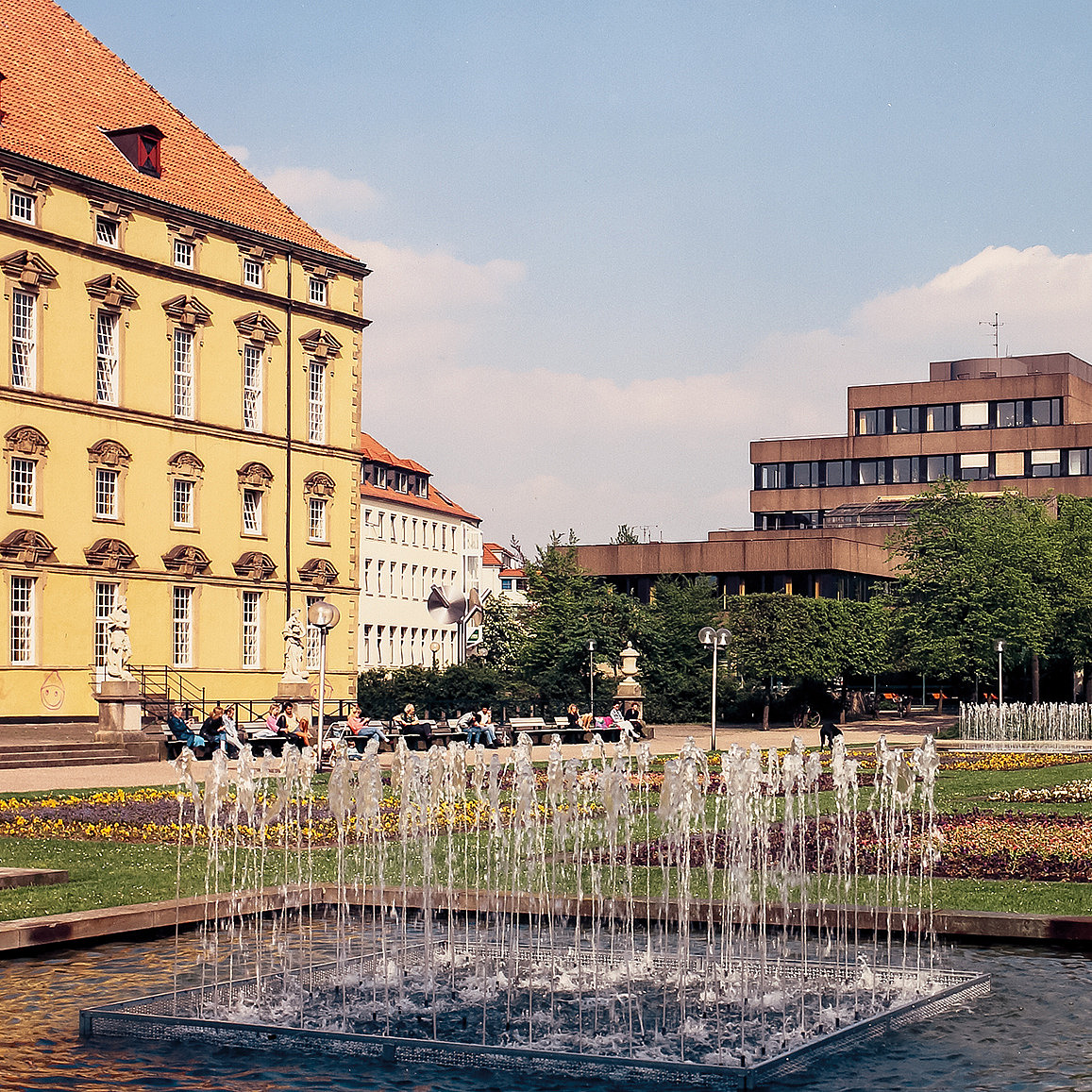 Color photo, taken in the castle garden. The castle front with the old terrace on the left edge of the picture. In the foreground are the fountains and flowerbeds that no longer exist, in the background the EW building with concrete front, without today's cladding.