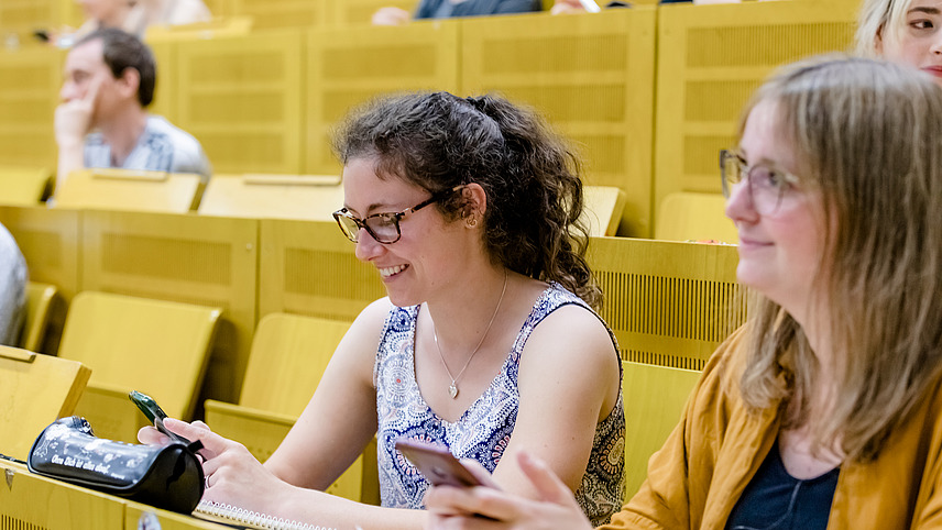 Studierende in Hörsaal, im Vordergrund zwei lächelnde junge Frauen