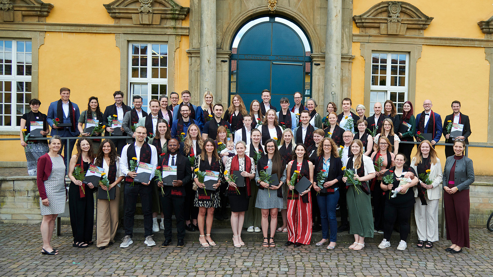 Eine Gruppe von Personen in festlicher Kleidung steht vor einem historischen Gebäude. Alle lächeln in die Kamera. Sie tragen alle einen eleganten Schal und halten eine Blume und eine Urkunde in der Hand.