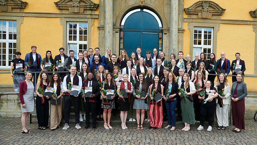Eine Gruppe von Personen in festlicher Kleidung steht vor einem historischen Gebäude. Alle lächeln in die Kamera. Sie tragen alle einen eleganten Schal und halten eine Blume und eine Urkunde in der Hand.