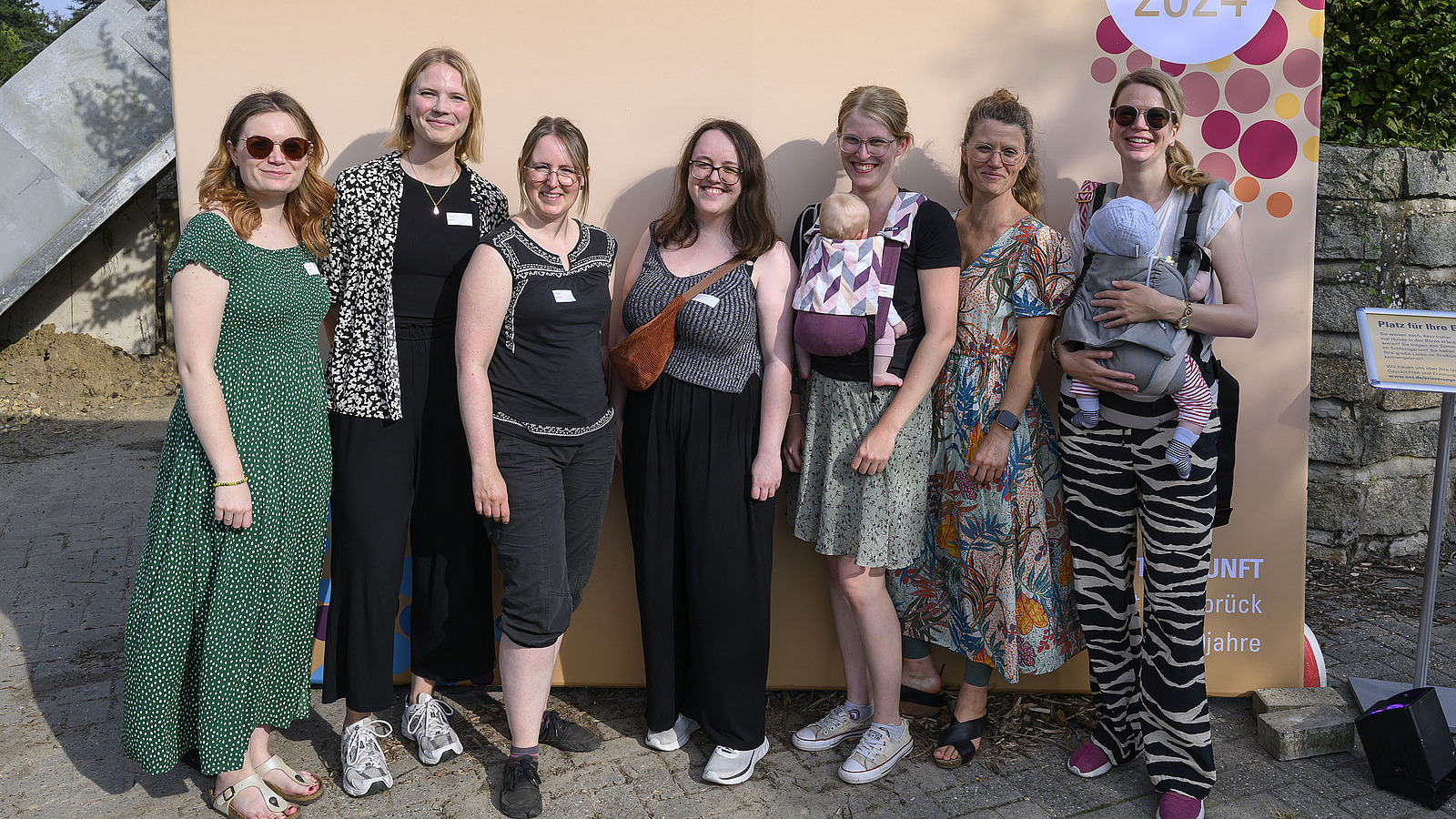 A group of people is standing in front of a photo wall, two women have baby carriers tied around them.