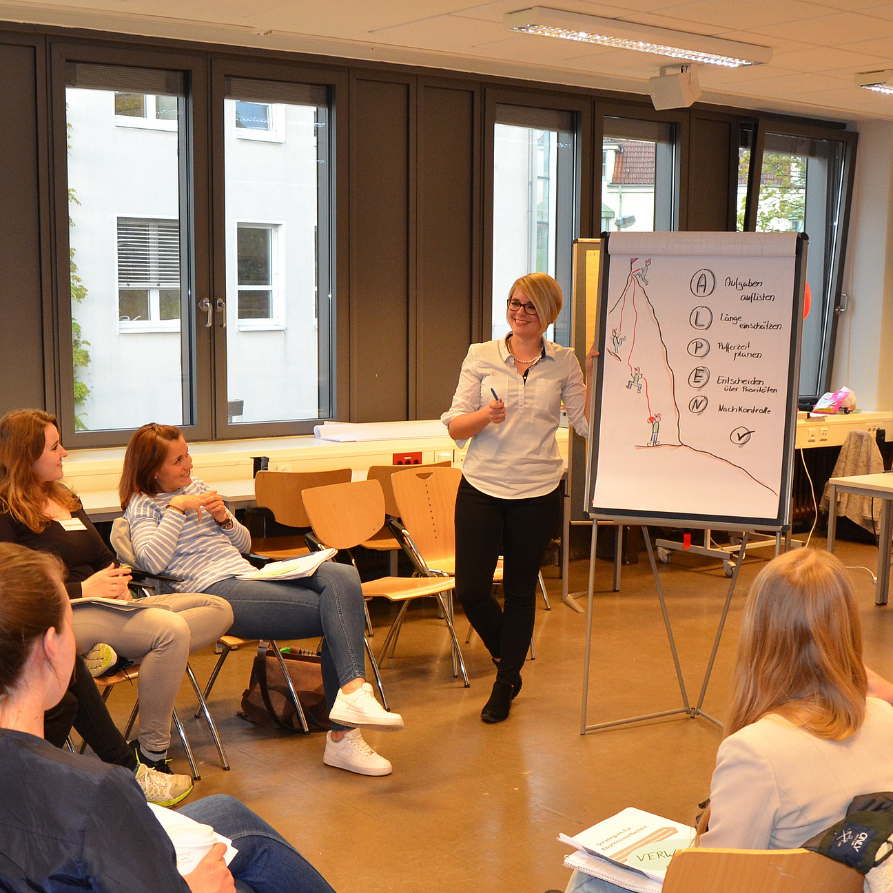 A lecturer stands at a flipchart. Students sit in a circle of chairs and listen.