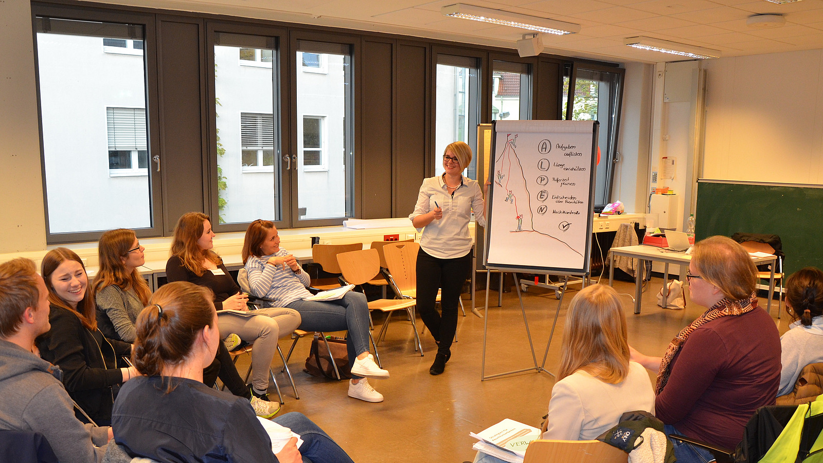 Eine Dozentin steht am Flipchart. Studierende sitzen im Stuhlkreis und hören zu. 