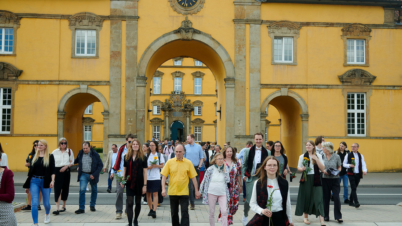 Zahlreiche Personen treten durch das Tor eines historischen Gebäudes, einige überqueren die davor liegende Straße