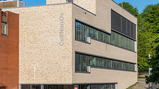 Das Bild zeigt ein modernes, dreistöckiges Gebäude mit einer hellen, strukturierten Fassade. An der Gebäudeseite ist der Schriftzug "CellNanOs" angebracht. Die Fassade weist viele große Fenster auf, die mit vertikalen Lamellen versehen sind. 