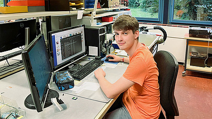Ein junger Mann sitzt an einem Schreibtisch in einem technischen Arbeitsraum. Vor ihm befindet sich ein Computer mit mehreren Bildschirmen, auf denen verschiedene Programme und Daten angezeigt werden. Der Schreibtisch ist mit technischen Geräten und Materialien gut bestückt.