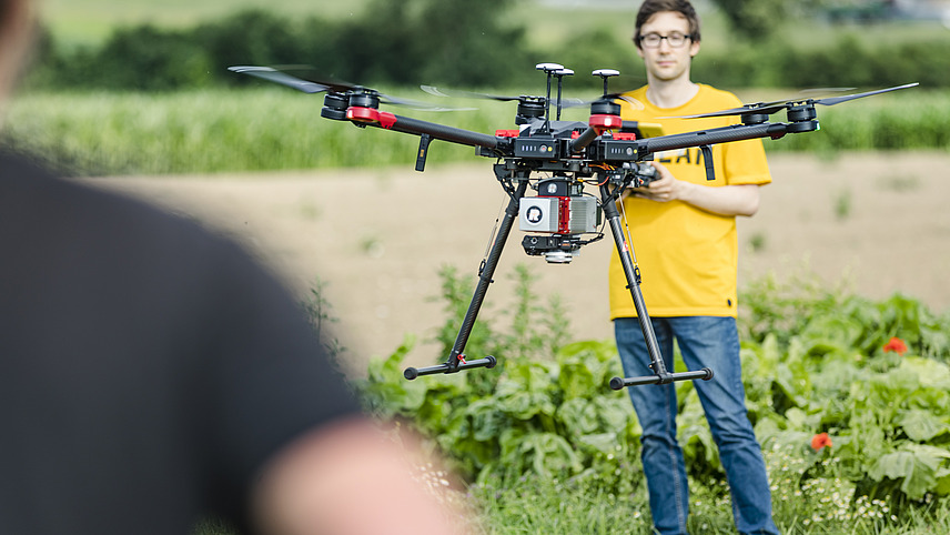 A flying drone can be seen from over a man's shoulder, controlled by another man. They are in a meadow in front of a field.