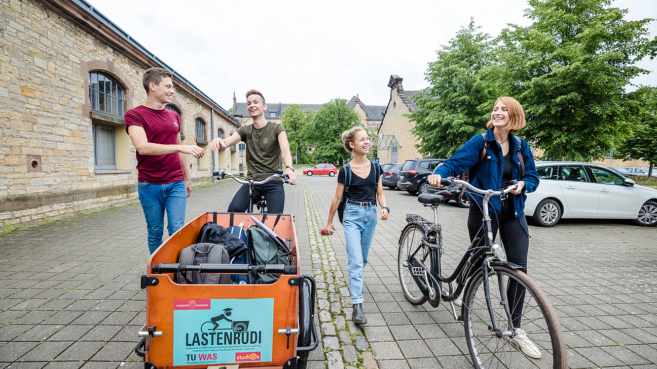 Vier Studierende. Zwei laufen zu Fuß, einer fährt ein Lastenrad, eine schiebt ihr Fahrrad.