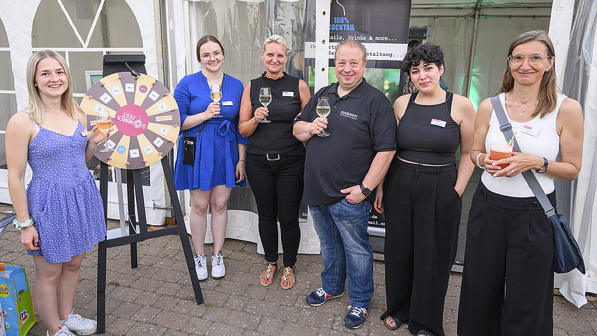 A group of people with drinks is standing next to a wheel of fortune.