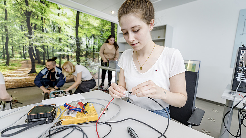 Ein paar Studierende hocken vor einem technischen Gerät auf dem Boden eines Werkraums. Eine Studentin im Vordergrund steckt ein schwarzes Kabel in ein kleines weißes Bauteil. Es liegen viele Kabel vor ihr.