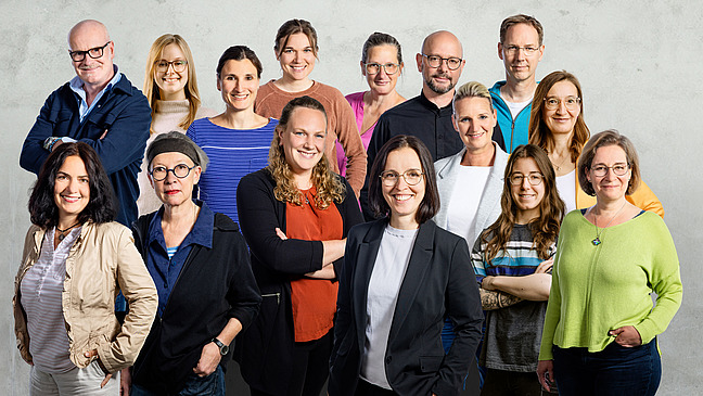 Eine Gruppe von 15 Personen steht in einer Gruppe zusammen, alle tragen legere unterschiedliche Kleidung.und blicken freundlich in die Kamera