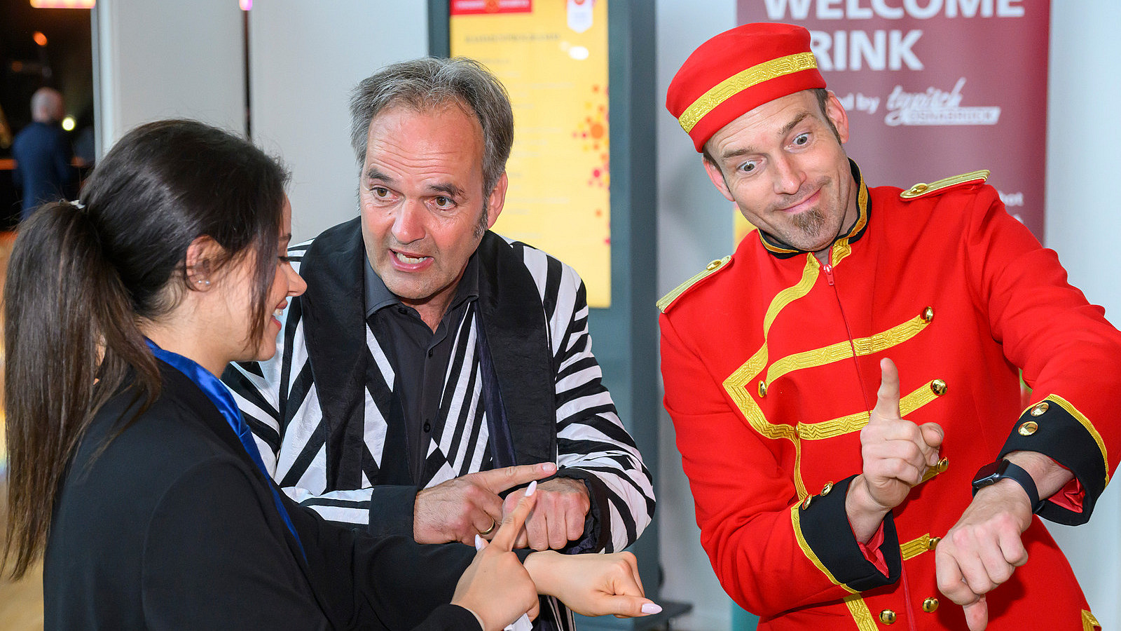 Three people, one of them in a bellboy costume, point their fingers at their wristwatches with an exaggerated gesture