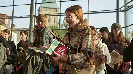 A group of people stands in a modern space with large windows. A young woman with glasses smiles while holding a colorful booklet. In the background, a church with a tall steeple is visible.