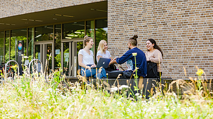 Four students sit on a bench in front of the library on Westerberg and talk.