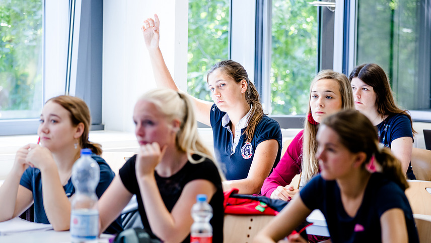 Sechs Studentinnen sitzen sommerlich gekleidet bei offenem Fenster in einem Seminarraum. Eine von ihnen hat die Hand gehoben