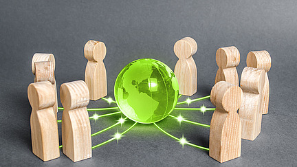 Holzfiguren stehen um eine leuchtend grüne, transparente Erdkugel, die in der Mitte platziert ist. Von der Kugel gehen grüne Linien aus, die eine Verbindung zwischen den Figuren symbolisieren.