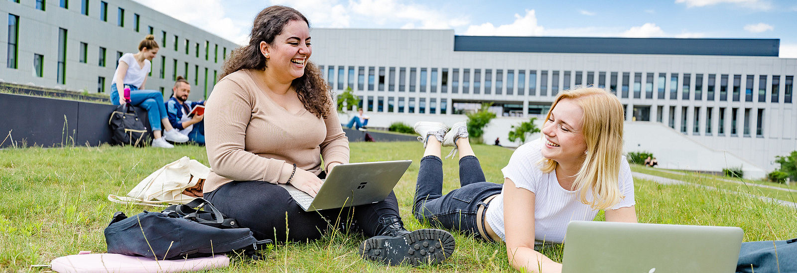 Zwei Studentinnen auf dem begrünten Platz neben der Bibliothek. Eine von ihnen sitzt, die andere liegt auf dem Rasen, beide haben Laptops vor sich