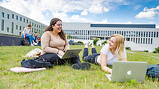 Zwei Studentinnen auf dem begrünten Platz neben der Bibliothek. Eine von ihnen sitzt, die andere liegt auf dem Rasen, beide haben Laptops vor sich