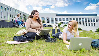 Zwei Studentinnen auf dem begrünten Platz neben der Bibliothek. Eine von ihnen sitzt, die andere liegt auf dem Rasen, beide haben Laptops vor sich