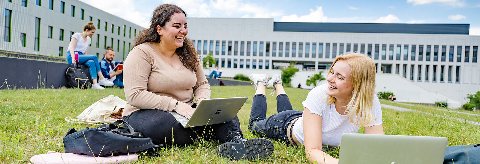 Zwei Studentinnen auf dem begrünten Platz neben der Bibliothek. Eine von ihnen sitzt, die andere liegt auf dem Rasen, beide haben Laptops vor sich