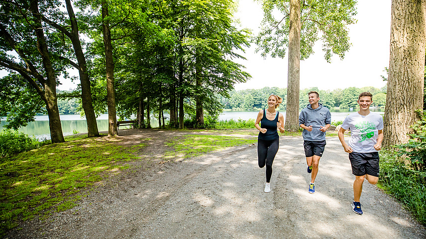 Drei Studierende joggen über einen asphaltierten Weg, der von Wiese und Bäumen gesäumt ist. Im Hintergrund ist der Rubbenbruchsee.