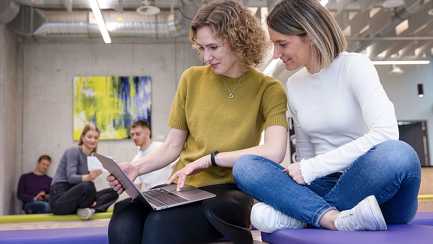 Two women sit next to each other on a colored seat and look at a laptop together. In the background, other people can be seen sitting together and communicating in a relaxed team environment