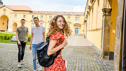 Eine Studentin im Schlossinnenhof schaut zurück zum Betrachter. Zwei Studenten gehen auf sie zu.