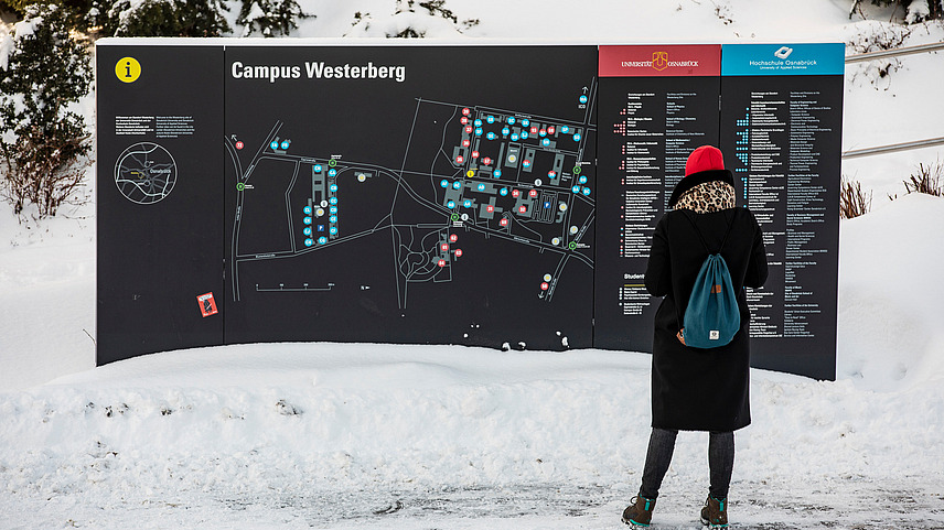 Eine Person mit Winterkleidung steht vor einer großen Wand mit dem Lageplan des Campus am Westerberg. Es liegt Schnee.