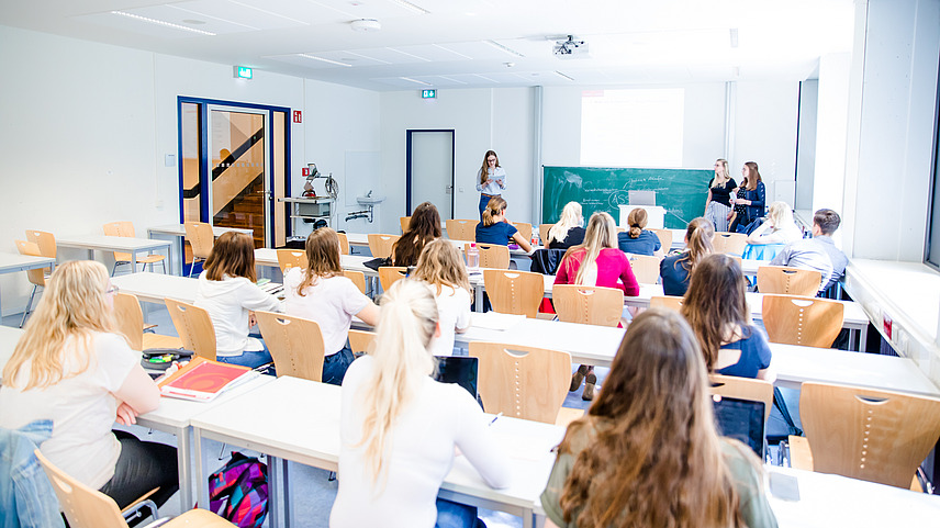 Blick von hinten über die Sitzreihen eines Seminarraums voller Studierender. Doktor Monika Voges leitet das Seminar und steht vorne neben zwei Studentinnen an der Tafel.